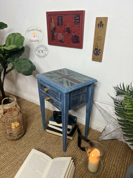Blue painted and distressed end table with large gold knobs on the 2 drawers. The London Vibe Transfer has been applied to the top, front, and side of the piece. Staged with a stack of books underneath it with a camera on top. The London Vibe Transfer has also been applied to some decorative pieces of wood that are hung on the wall.