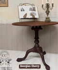 A small, round wooden table with a detailed, ornate pedestal base is displayed. On top are an open book on a stand and a decorative Victorian-style candelabra. A logo and the words "Georgian Cherry" are visible at the bottom.