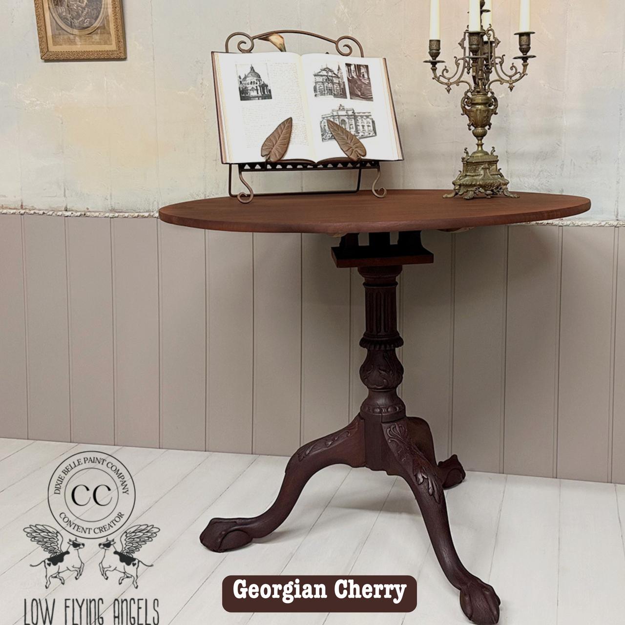 A small, round wooden table with a detailed, ornate pedestal base is displayed. On top are an open book on a stand and a decorative Victorian-style candelabra. A logo and the words "Georgian Cherry" are visible at the bottom.