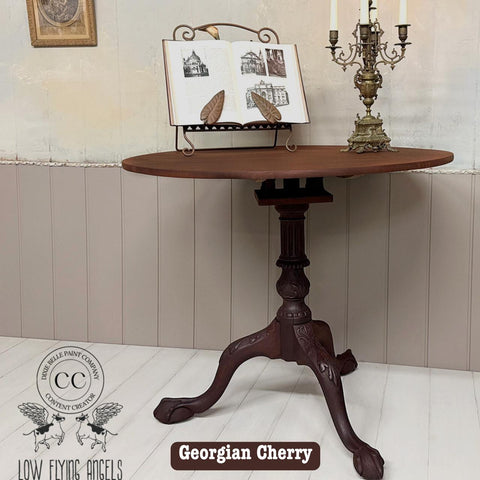 A small, round wooden table with a detailed, ornate pedestal base is displayed. On top are an open book on a stand and a decorative Victorian-style candelabra. A logo and the words "Georgian Cherry" are visible at the bottom.