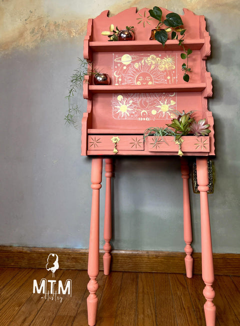 Pink painted tall shelf with the Lunar Solstice Transfer applied on the middle back of the piece and outlined in gold. Staged with faux succulents in one of the drawers and small pots with plants hanging over the edge on the shelves.