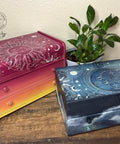 Two wooden boxes with celestial designs sit on a wooden surface. The left box is reddish pink and yellow with a sun motif. The right box is blue with a moon and stars design. A green plant is nearby. A logo in the top left corner reads "Dixie Belle Paint Company Content Creator."
