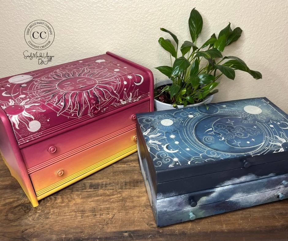 Two wooden boxes with celestial designs sit on a wooden surface. The left box is reddish pink and yellow with a sun motif. The right box is blue with a moon and stars design. A green plant is nearby. A logo in the top left corner reads "Dixie Belle Paint Company Content Creator."