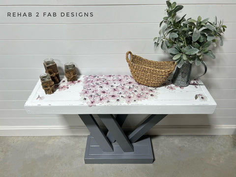 A white and gray wooden table with a crisscross base is decorated with a metal watering can full of green foliage, a woven basket, and three candles. Lilac flowers are on the table's white surface using the Magnificent Magnolia Transfer. The words "REHAB 2 FAB DESIGNS" are on the wall above.