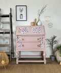 A vintage pink secretary desk adorned with floral designs is positioned against a white wall. It is flanked by a decorative wooden ladder with fairy lights on the left, a framed botanical print above, potted plants, and a wicker lamp on the floor.