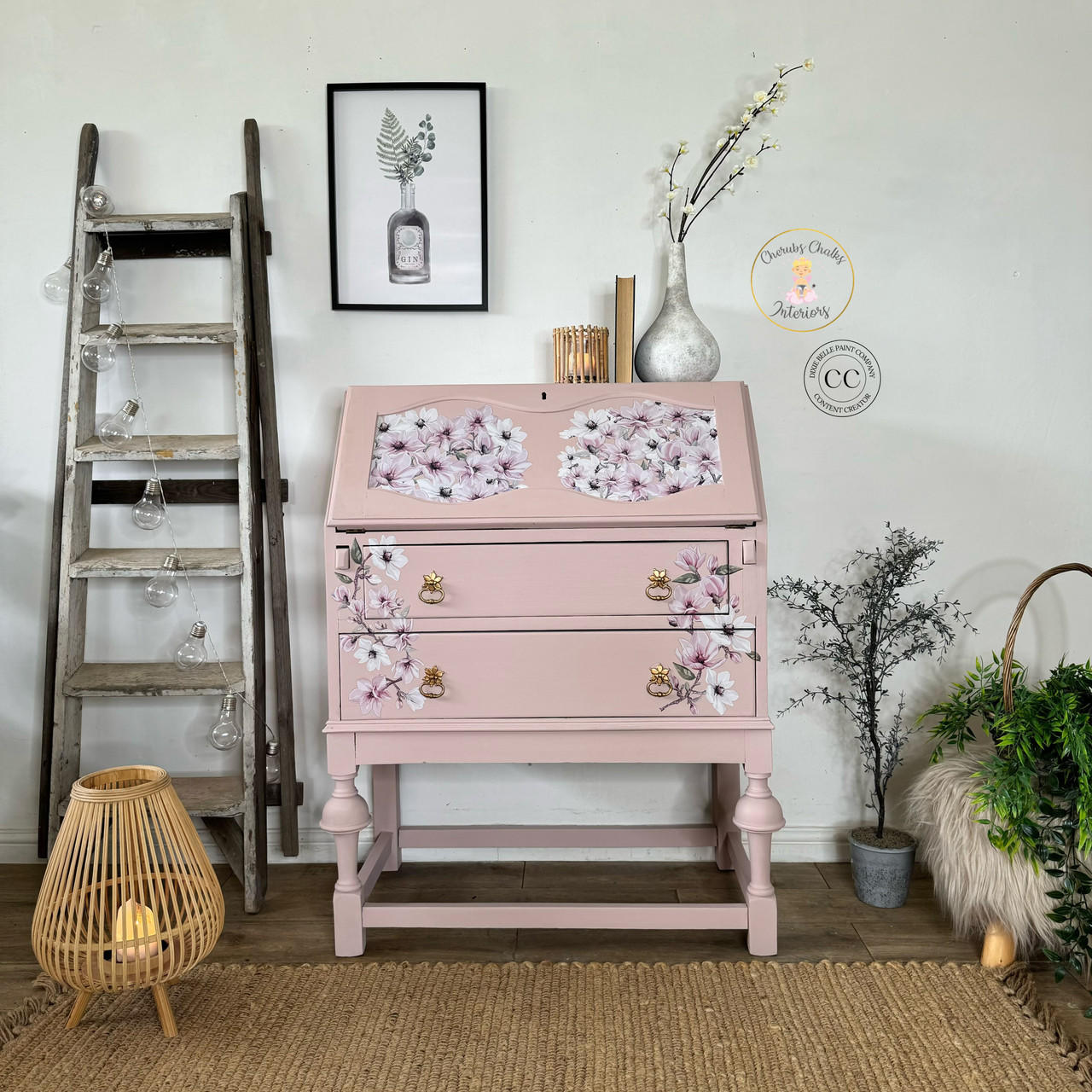 A vintage pink secretary desk adorned with floral designs is positioned against a white wall. It is flanked by a decorative wooden ladder with fairy lights on the left, a framed botanical print above, potted plants, and a wicker lamp on the floor.
