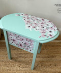 A small oval-shaped table with light turquoise paint and a pattern of pink and white flowers on the surface and side. The table is placed on a woven rug and there is a basket visible in the background. The table has a delicate, artistic design by Faff Designs.