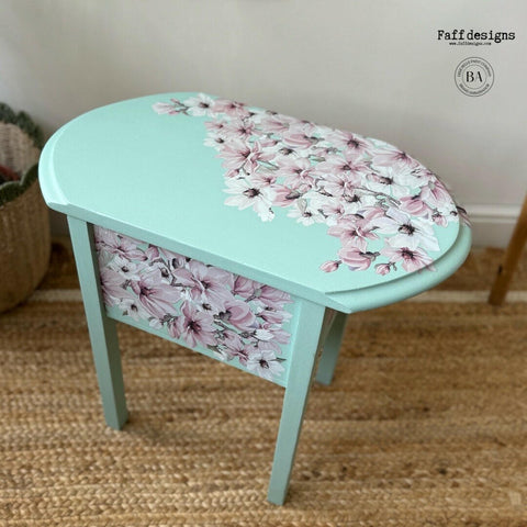 A small oval-shaped table with light turquoise paint and a pattern of pink and white flowers on the surface and side. The table is placed on a woven rug and there is a basket visible in the background. The table has a delicate, artistic design by Faff Designs.