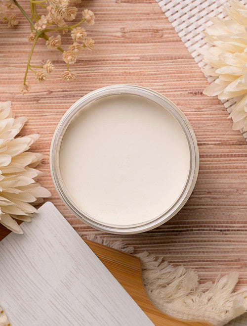 An open jar of cream-colored paint on a wooden surface, surrounded by dried flowers and textured fabric accents. The cream appears smooth and white, with a rustic and natural aesthetic.