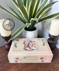A decorative wooden box with floral and cowboy boots design sits on a wooden table. It is flanked by two candles in dark brown holders. A potted plant with long, variegated leaves is in the background. A logo in the top left corner reads "AJ's Vintage Designs.