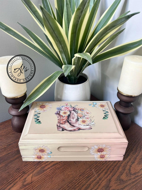 A decorative wooden box with floral and cowboy boots design sits on a wooden table. It is flanked by two candles in dark brown holders. A potted plant with long, variegated leaves is in the background. A logo in the top left corner reads "AJ's Vintage Designs.