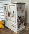 A white, vintage-style jewelry cabinet with floral and horse-themed designs is displayed. The cabinet features intricate handles, horsehead artwork on the side, and floral decals on the doors. A bright yellow sunflower lies beside it on a wooden surface.