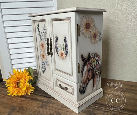 A white, vintage-style jewelry cabinet with floral and horse-themed designs is displayed. The cabinet features intricate handles, horsehead artwork on the side, and floral decals on the doors. A bright yellow sunflower lies beside it on a wooden surface.