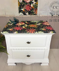 A white two-drawer nightstand with a colorful, floral-patterned top sits on a wooden floor. A small bird figurine and a green fern decorate the scene. A matching floral artwork hangs on the wall behind.
