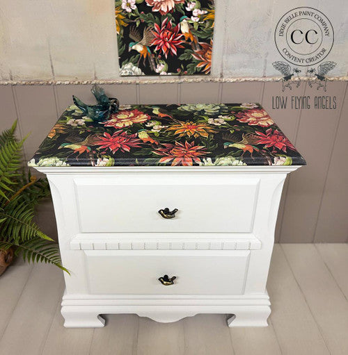 A white two-drawer nightstand with a colorful, floral-patterned top sits on a wooden floor. A small bird figurine and a green fern decorate the scene. A matching floral artwork hangs on the wall behind.