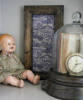 A vintage doll with a worn-out facial expression and body sits next to a framed toile-patterned picture and under a glass dome containing a pocket watch and book on a shelf.