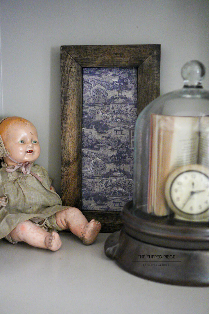 A vintage doll with a worn-out facial expression and body sits next to a framed toile-patterned picture and under a glass dome containing a pocket watch and book on a shelf.