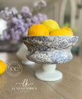 A blue and white ornate bowl on a pedestal filled with bright yellow lemons, placed on a wooden table. In the background, there is a blurred bouquet of lilac flowers. The text "Leah Kendrick Design" and "Dixie Belle Paint Company Content Creator" appears at the bottom.