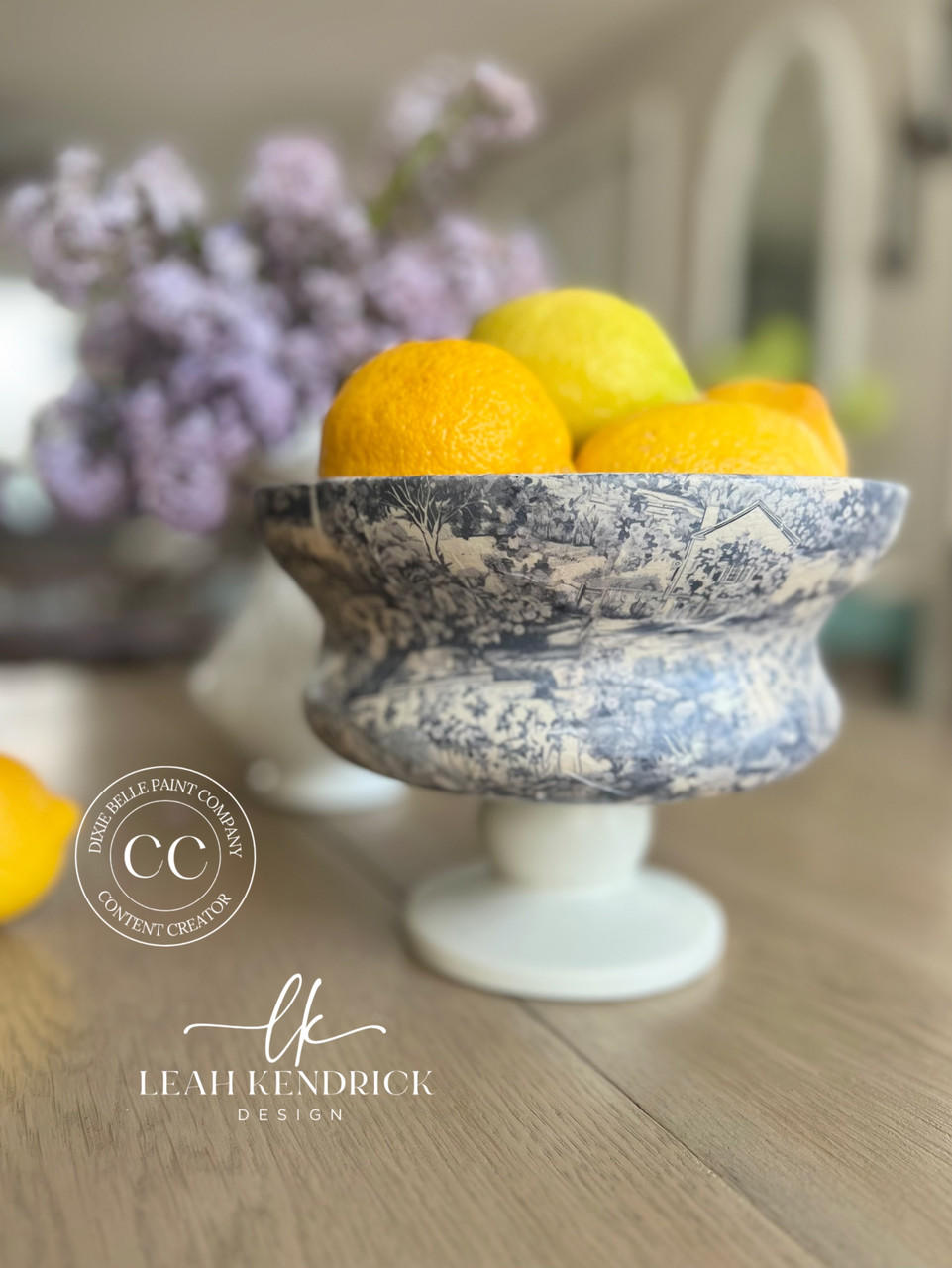 A blue and white ornate bowl on a pedestal filled with bright yellow lemons, placed on a wooden table. In the background, there is a blurred bouquet of lilac flowers. The text "Leah Kendrick Design" and "Dixie Belle Paint Company Content Creator" appears at the bottom.