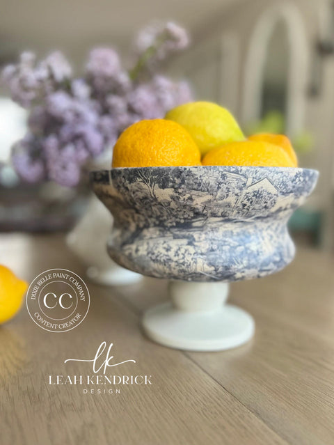 A blue and white ornate bowl on a pedestal filled with bright yellow lemons, placed on a wooden table. In the background, there is a blurred bouquet of lilac flowers. The text "Leah Kendrick Design" and "Dixie Belle Paint Company Content Creator" appears at the bottom.