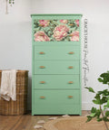 A mint green dresser with four drawers, the top drawer decorated with a floral decoupage paper featuring large pink flowers. The dresser stands on a wooden floor, surrounded by plants and a wicker basket.