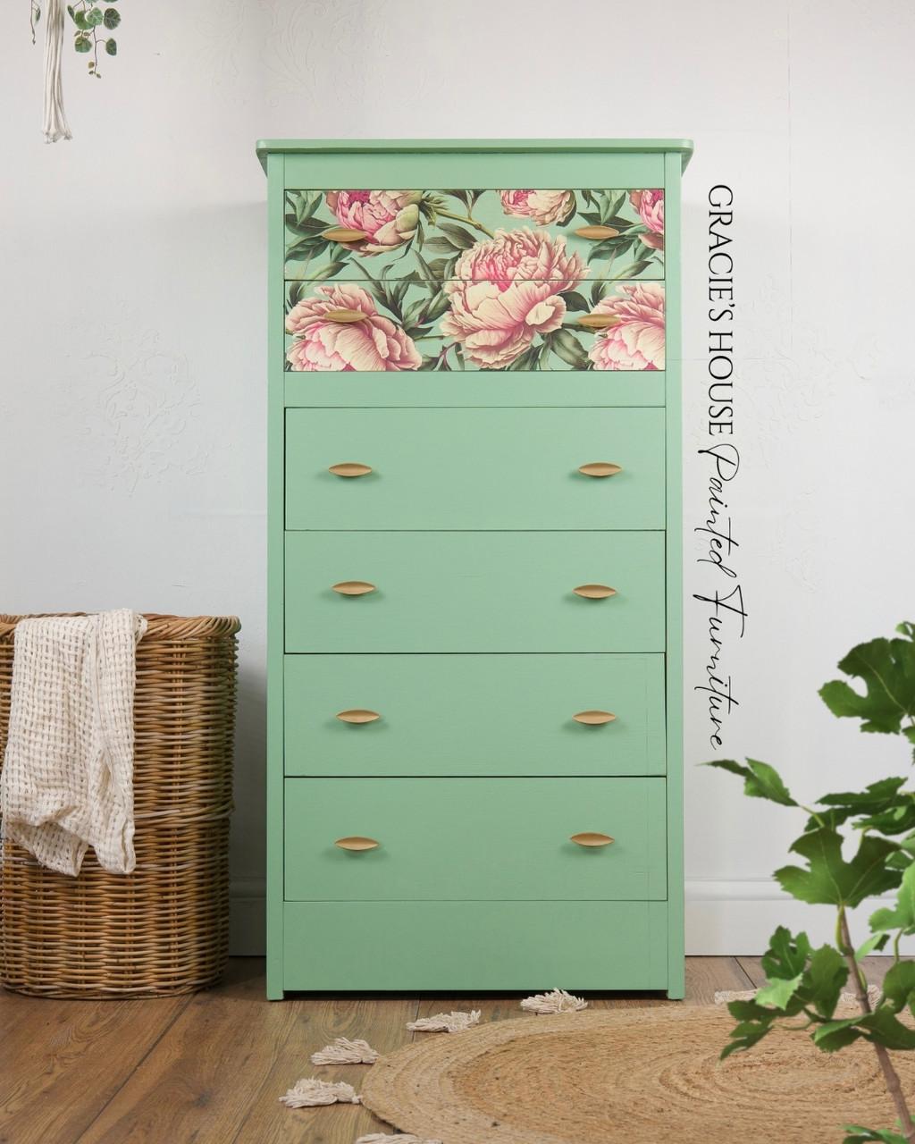 A mint green dresser with four drawers, the top drawer decorated with a floral decoupage paper featuring large pink flowers. The dresser stands on a wooden floor, surrounded by plants and a wicker basket.