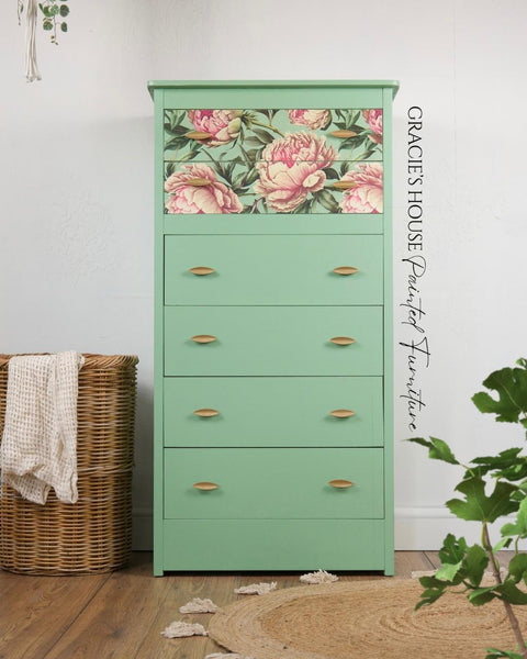 A mint green dresser with four drawers, the top drawer decorated with a floral decoupage paper featuring large pink flowers. The dresser stands on a wooden floor, surrounded by plants and a wicker basket.