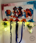 A colorful wall-mounted rack featuring a vibrant painting of a dog and paw prints, with three hooks holding leashes. Two glass jars with fairy lights and white flowers hang from the ends of the rack, adding a decorative touch.