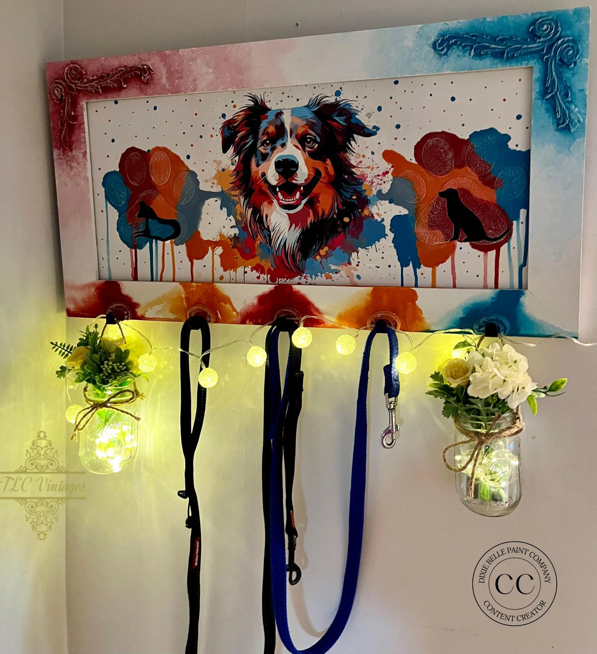 A colorful wall-mounted rack featuring a vibrant painting of a dog and paw prints, with three hooks holding leashes. Two glass jars with fairy lights and white flowers hang from the ends of the rack, adding a decorative touch.