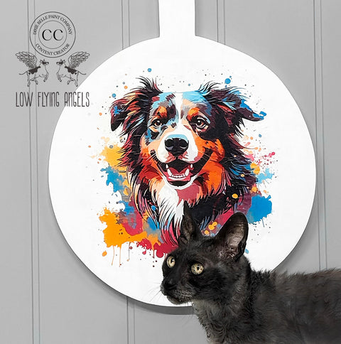 A colorful painted image of a happy dog is displayed on a circular white background mounted on a wall. A black cat sits in the foreground, looking upwards. The text "Low Flying Angels" is visible on the left side of the image, alongside a logo of flying cows.