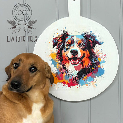 A tan and white dog is sitting in front of a painted wooden paddle featuring a colorful, abstract painting of another dog. The backdrop has a logo with the text "LOW FLYING ANGELS" and a circular emblem.