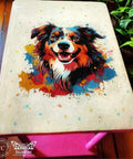 A colorful painting of a dog with a happy expression on a table surface, surrounded by vibrant splashes of paint. The dog's detailed fur is in shades of brown, black, and white.