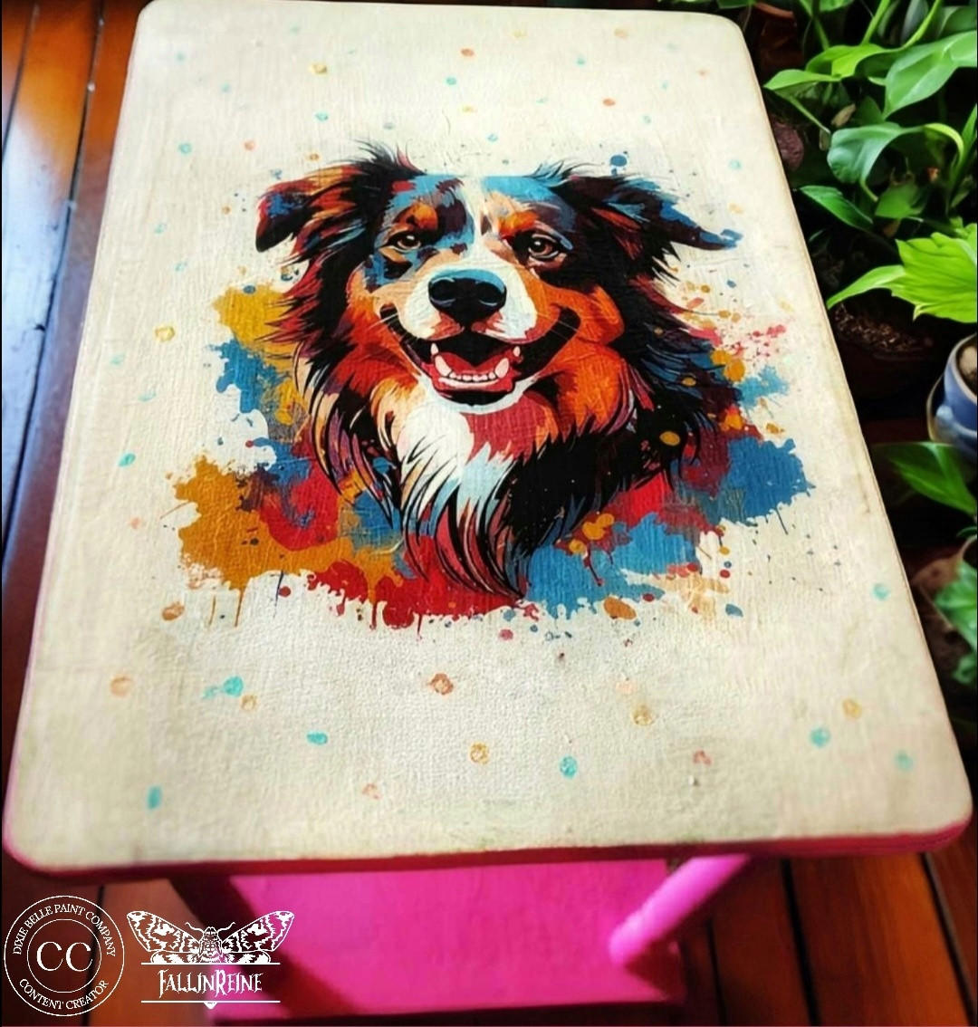 A colorful painting of a dog with a happy expression on a table surface, surrounded by vibrant splashes of paint. The dog's detailed fur is in shades of brown, black, and white.