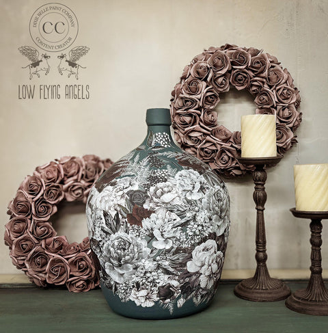 A large, intricately decorated ceramic floral vase is centered against a light wall. It is flanked by two metal candlesticks with candles and adorned with two wreaths made of dusty rose-colored flowers. The backdrop features a logo for "Low Flying Angels."