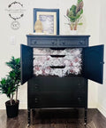 A refurbished vintage black dresser with floral patterned drawers and doors open, with the Monochrome Majesty Transfer applied on the inside drawers. A framed picture and plants are on top, and a large potted plant is on the side, in a bright room.