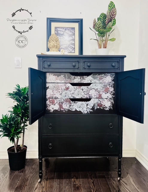 A refurbished vintage black dresser with floral patterned drawers and doors open, with the Monochrome Majesty Transfer applied on the inside drawers. A framed picture and plants are on top, and a large potted plant is on the side, in a bright room.