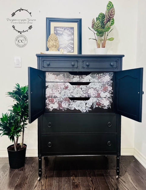 A refurbished vintage black dresser with floral patterned drawers and doors open, with the Monochrome Majesty Transfer applied on the inside drawers. A framed picture and plants are on top, and a large potted plant is on the side, in a bright room.