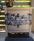 A cream-colored painted wooden nightstand with Monochrome Majesty Transfer applied on the drawers, standing on a porch with a plant pot on top, under a sunny and shaded backdrop.