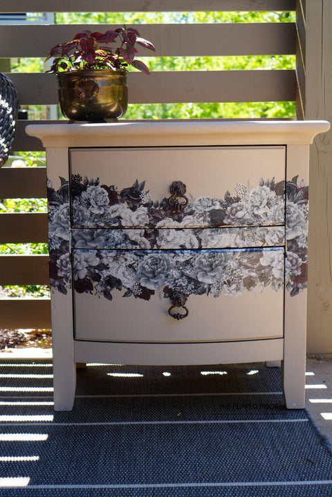 A cream-colored painted wooden nightstand with Monochrome Majesty Transfer applied on the drawers, standing on a porch with a plant pot on top, under a sunny and shaded backdrop.