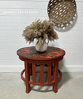 A round wooden end table with a red and black patterned top using the Moody Mums A1 Rice Decoupage Paper. Adorned with a white vase filled with fluffy pampas grass, set against a white shiplap wall with a decorative round wooden tray hanging.
