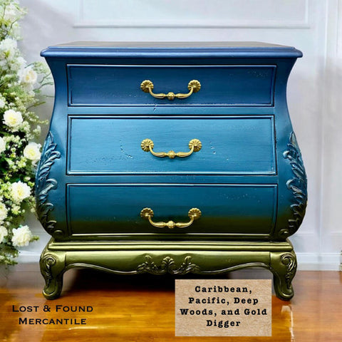 A vintage chest of drawers painted in blue ombre with gold accents and ornate handles, sits on a wooden floor next to white flowers. Label reads “Caribbean, Pacific, Deep Woods, and Gold Digger.”.
