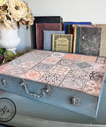 A vintage-style box with a mosaic tile design sits on a table. Behind it, there is a collection of old books and a ceramic pot with dried flowers. The box has a handle and metal clasps. Logo at the bottom reads "AJ's Vintage Designs."