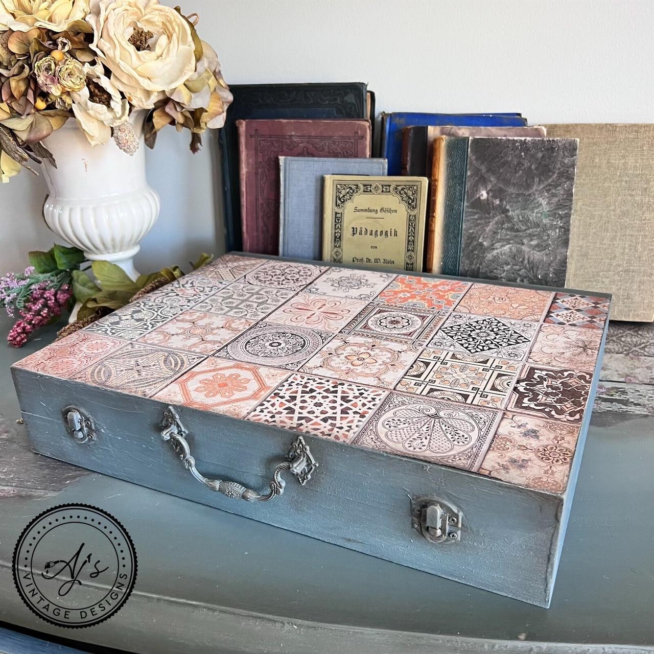 A vintage-style box with a mosaic tile design sits on a table. Behind it, there is a collection of old books and a ceramic pot with dried flowers. The box has a handle and metal clasps. Logo at the bottom reads "AJ's Vintage Designs."
