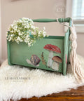 Green chalk painted magazine holder with Mr. Toad's Transfer applied on one of the sides. Staged with white flowers inside and macrame hanging off one of the sides.
