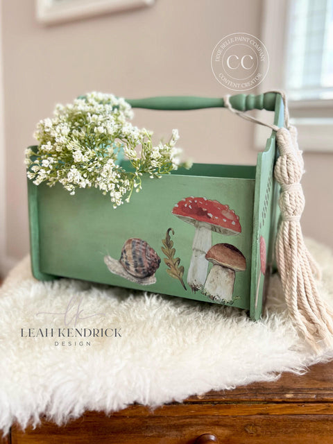 Green chalk painted magazine holder with Mr. Toad's Transfer applied on one of the sides. Staged with white flowers inside and macrame hanging off one of the sides.