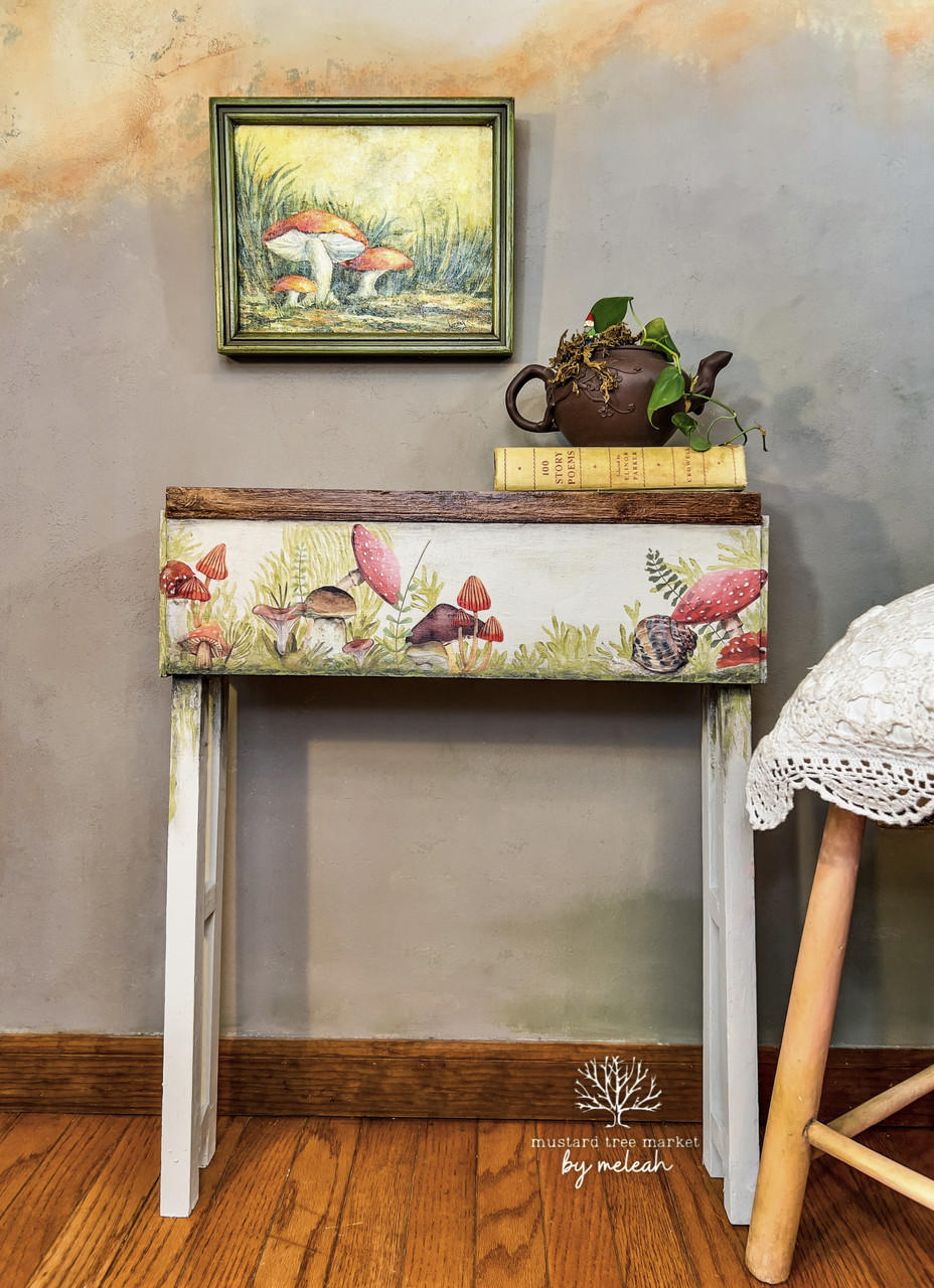 White chalk painted table with a dark wood top. Mr. Toad's Transfer has been applied on the front of the table. Staged with a book and a metal watering can, with a pothos inside, on top.