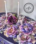 A table adorned with vibrant, large floral patterns in shades of purple, pink, and blue. Two white candles in gold holders and a framed photograph of a woman are placed on the table. A logo in the corner reads "AJ's Vintage Designs.