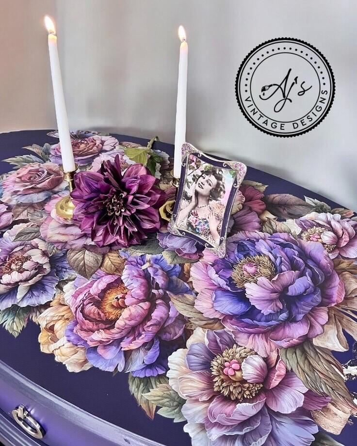 A table adorned with vibrant, large floral patterns in shades of purple, pink, and blue. Two white candles in gold holders and a framed photograph of a woman are placed on the table. A logo in the corner reads "AJ's Vintage Designs.