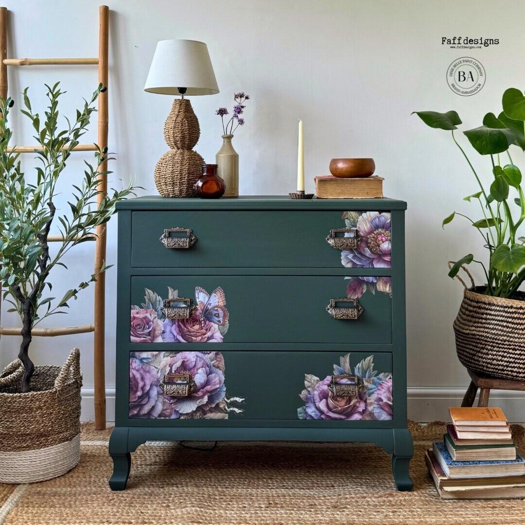 A vibrant, nature-themed room features a dark green dresser adorned with detailed floral and butterfly decals. The dresser is flanked by plants in woven baskets and accented with a lamp, vases, and decorative items on top. A wooden ladder stands to the left.