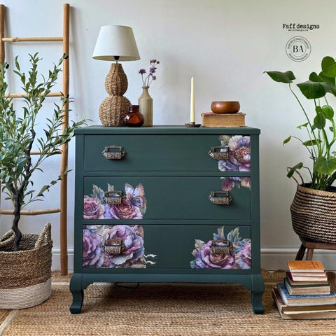A vibrant, nature-themed room features a dark green dresser adorned with detailed floral and butterfly decals. The dresser is flanked by plants in woven baskets and accented with a lamp, vases, and decorative items on top. A wooden ladder stands to the left.